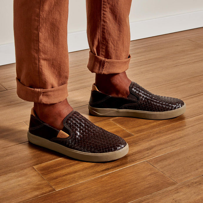 Dark brown OluKai Lae'Ahi woven leather slip-ons worn with rolled trousers on wooden floor