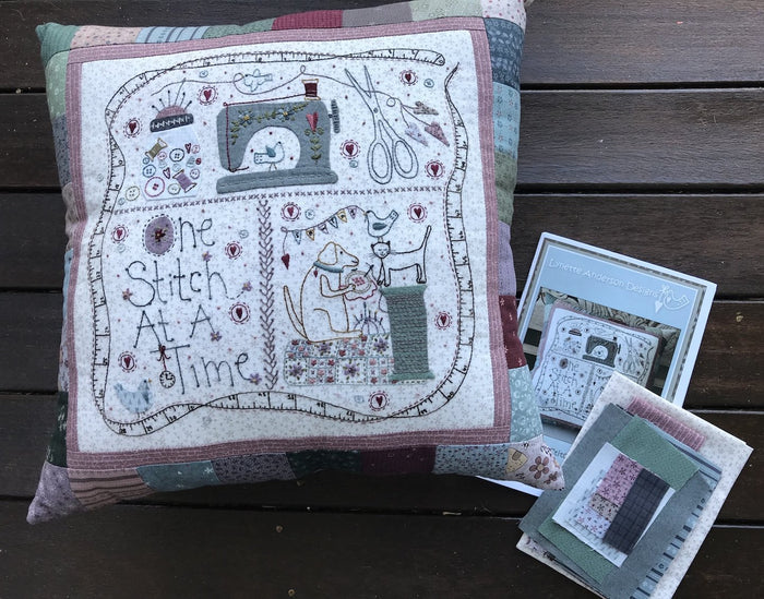 A pillow kit with a color pattern and embroidery design, titled 'The Stitch at a Time', displayed on a wooden surface.