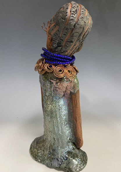 Meet Ophelia! Ophelia stands 7.5" x 4" x 5" and weighs 1.02  lbs. She has a lovely honey brown complexion with reddish brown lips. She has a short braided hairstyle.  Ophelia has a colorful metallic antique copper glazed dress. She wears spiral copper wire necklaces on top of an aqua blue beaded collar. Ophelia long loving arms rest at her side. With accent blue eye shadow and eyes wide opened, Ophelia has hopes of finding  her way into your home!