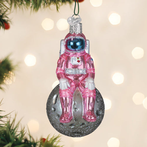 Pink astronaut glass ornament seated on the moon hanging on a tree