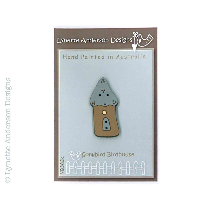 A hand painted wooden button with a blue roof on a grey background, labeled with the artist's signature and the brand name Songbird Birdhouse.
